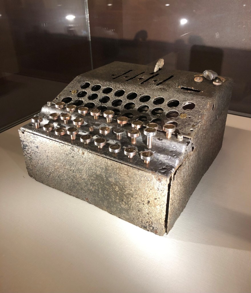 Picture shows a colour photo of a silver grey coloured metal device with a mechanical keyboard. This is the Enigma G 110, or 'counter' Enigma, a cipher machine used by the German military to encipher messages during the Second World War. This machine is at Bletchley Park, which was the centre of Allied code breaking operations in the war.