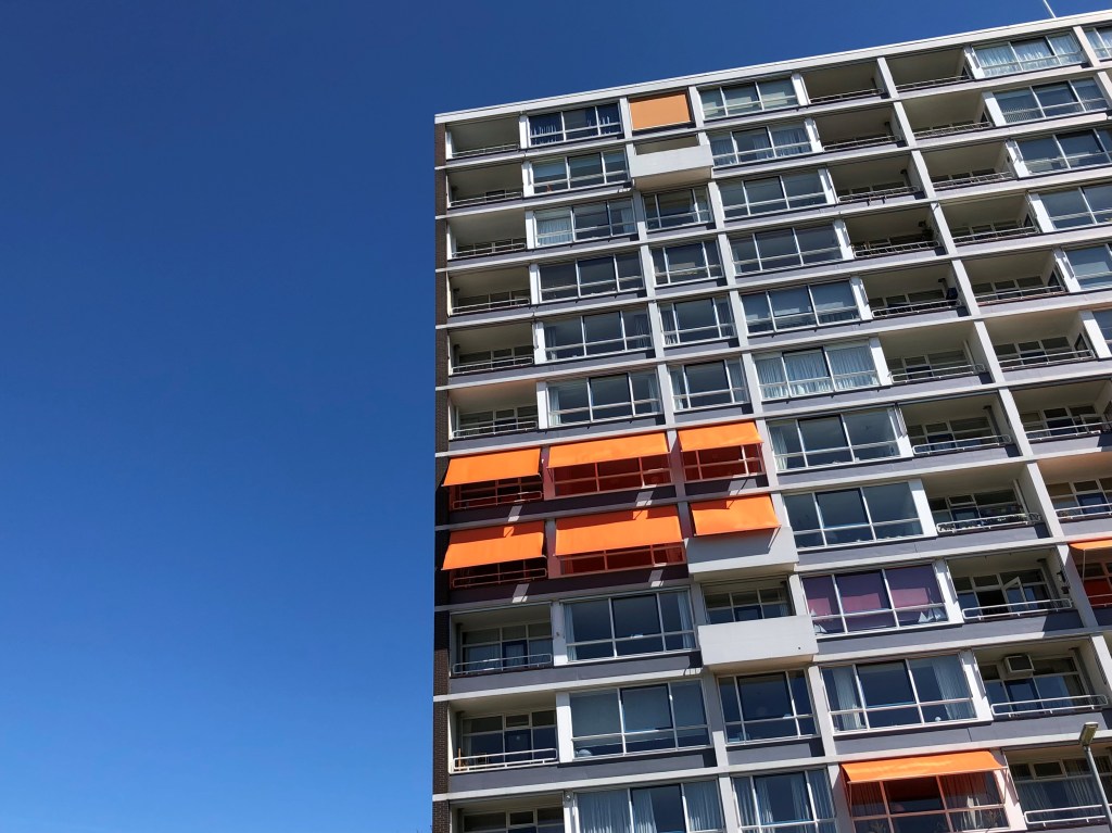 Picture shows a colour photo of a multi storey apartment block, 11 stories are visible, against a bright blue summer sky. Some of the apartments have bright orange sun shades over the windows. Location is Rotterdam in the Netherlands.
