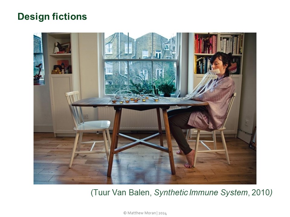 Design fiction by Tuur Van Balen entitled Synthetic Immune System, from 2010. In the picture person sits at a table in a domestic interior. A number of tubes extend from the persons mouth and are connected to smal glass balls on the table. The work was designed to accompany research testing the externalisation of our immune system by delegating certain metabolisation processes to outsourced micro-organisms.