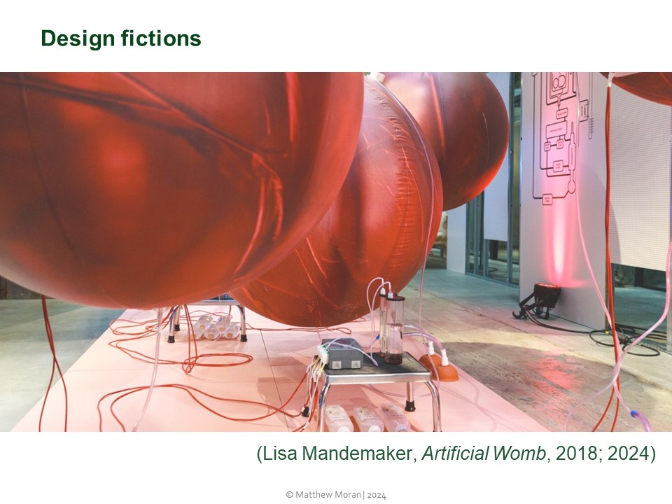 Picture shows a design fiction entitled Artifical Womb by Lisa Mandemaker, on display at Dutch Design Week in 2018. The work consists of four large floating red ballons connected by thin red cables to each other and to a small grey pump device and glass cylinders on a grey steel stool.
