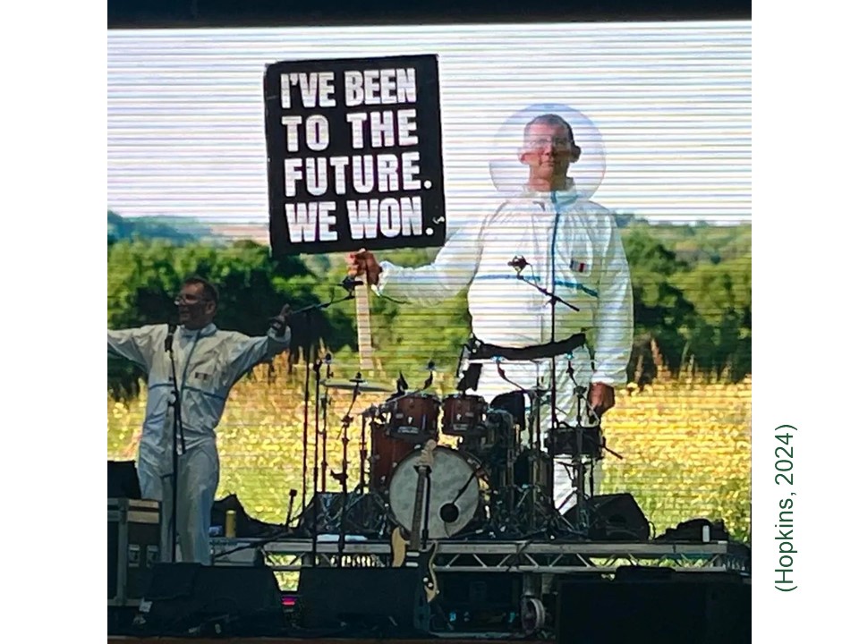 Picture shows the author and activist Rob Hopkins on stage at a music festival wearing an astronaut suit (without helmet), arms raised addressing the crowd. Behind him, a huge digital screen (seen behind a large drum kit) displays a picture of Hopkins still in the astronaut suit, now with helmet on, standing in a field and holding a placard on which is written I've been to the future. We won. 
