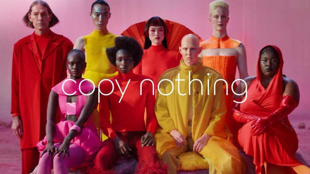 Picture is a still from a TV advert from the Jaguar cars launch of the Type 00 car, showing two rows of four people (four standing, four sitting), wearing bright yellow, pink and red clothes, in a vibrant pink setting, and the words copy nothing superimposed.