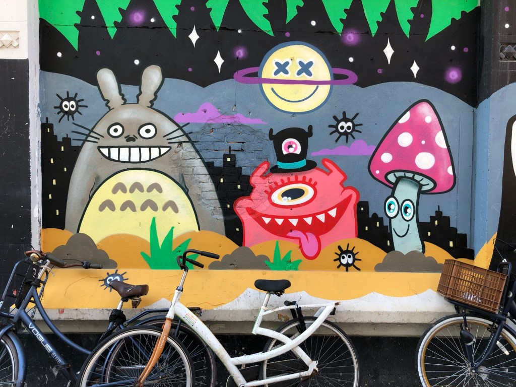 Picture is a colour photo of street art in Rotterdam, the Netherlands, showing stylised figures of a grey rabbit, a nondescript pink one-eyed monster with a second eye in its top hat, and a rather nerdy looking toadstool.